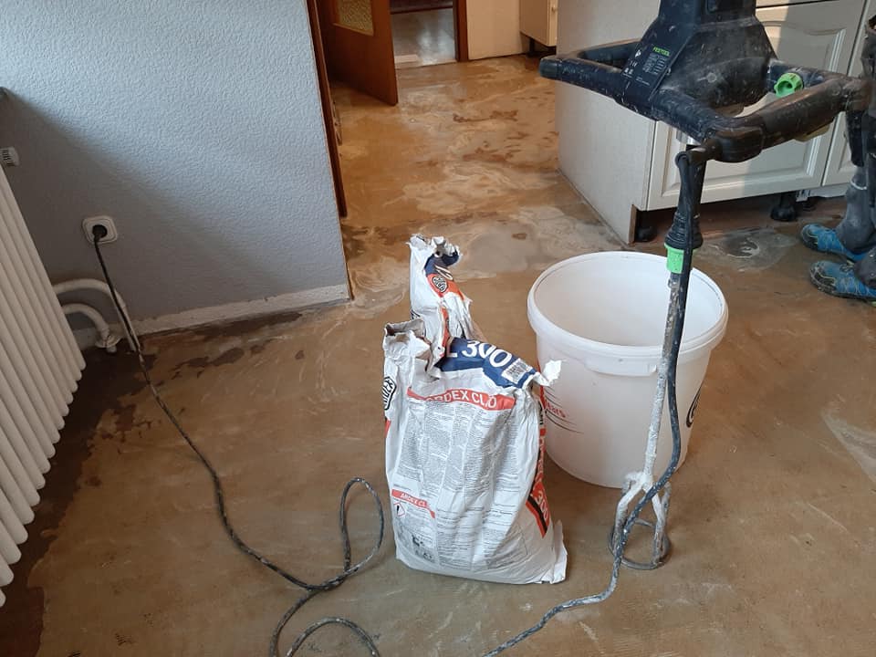 Materialien und Werkzeuge für ein Hausrenovierungsprojekt, darunter Bodenverlegungsmaterialien, ein teilweise verbrauchter Beutel mit Spachtelmasse und ein Mixer, liegen auf dem Boden verstreut. Fussbodentechnik Schwallach -Bodenbeläge, Vinylbelag, Parkett - Neckaraue 7 - 71686 Remseck