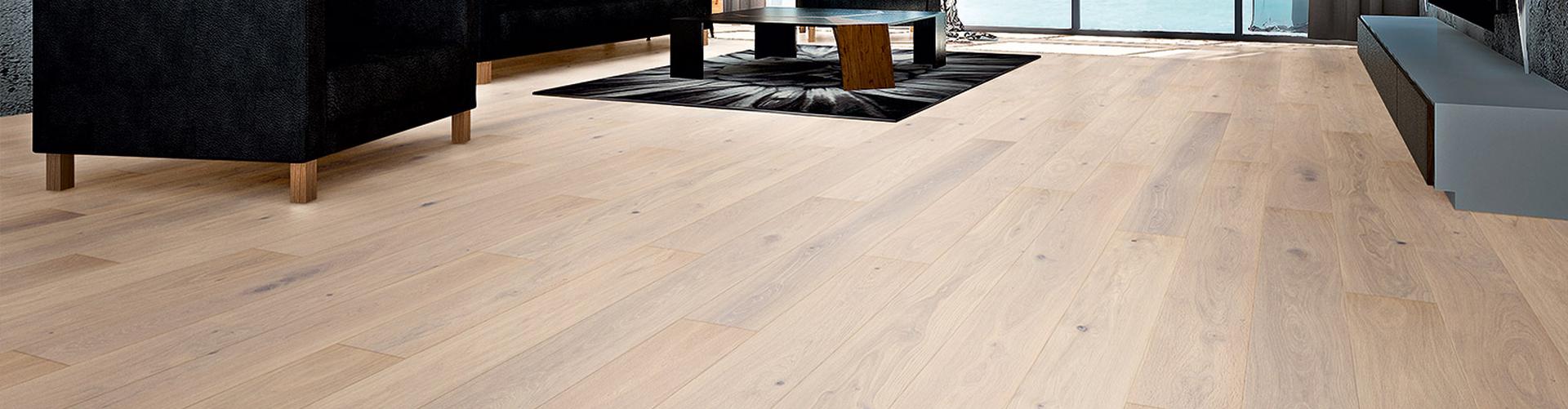 Ein modernes Wohnzimmer im aktuellen Design mit hellem Holzboden, einem schwarzen Sofa, einem runden Couchtisch und einem Teppich. Große Fenster durchfluten den Raum mit reichlich natürlichem Licht. - Fussbodentechnik Schwallach -Bodenbeläge, Vinylbelag, Parkett - Neckaraue 7 - 71686 Remseck
