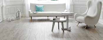 Ein modernes Wohnzimmer mit einem weißen Sofa, einem weißen Sessel, einem kleinen runden Tisch und einem Beistelltisch. Der Boden ist aus Holz und auf dem Sofa liegt ein dekoratives Kissen. - Fussbodentechnik Schwallach -Bodenbeläge, Vinylbelag, Parkett - Neckaraue 7 - 71686 Remseck