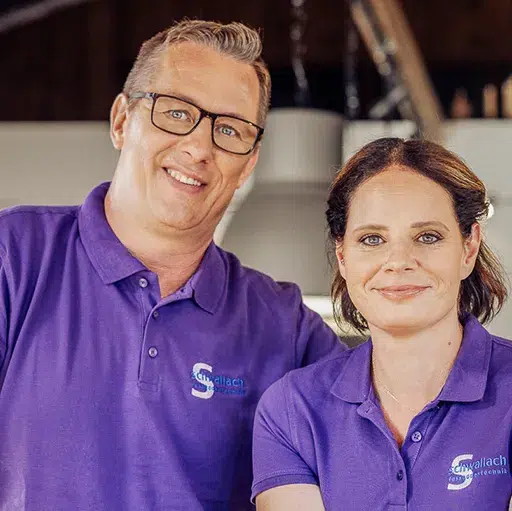 Alexandra und Michael Schwallach in passenden lila Kontakt-Shirts mit einem Logo stehen drinnen nebeneinander und lächeln in die Kamera. - Fussbodentechnik Schwallach -Bodenbeläge, Vinylbelag, Parkett - Neckaraue 7 - 71686 Remseck