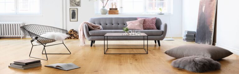 Ein modernes Wohnzimmer mit einem grauen Sofa, einem Metallstuhl, einem quadratischen Couchtisch und dekorativen Kissen. Bücher und eine Felldecke liegen auf dem Holzboden. Große Fenster lassen Tageslicht herein. - Fussbodentechnik Schwallach -Bodenbeläge, Vinylbelag, Parkett - Neckaraue 7 - 71686 Remseck