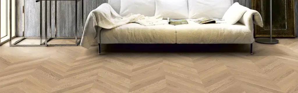 Ein weißes Sofa mit einer Decke und einem Buch steht auf einem Holzboden mit Fischgrätenmuster und präsentiert Produkte in einem hellen Raum. - Fussbodentechnik Schwallach -Bodenbeläge, Vinylbelag, Parkett - Neckaraue 7 - 71686 Remseck